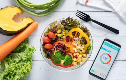 Eine Schüssel mit sehr viel frischem Gemüse steht auf einem Tisch, daneben ein Handy mit einer Abnehm-App.