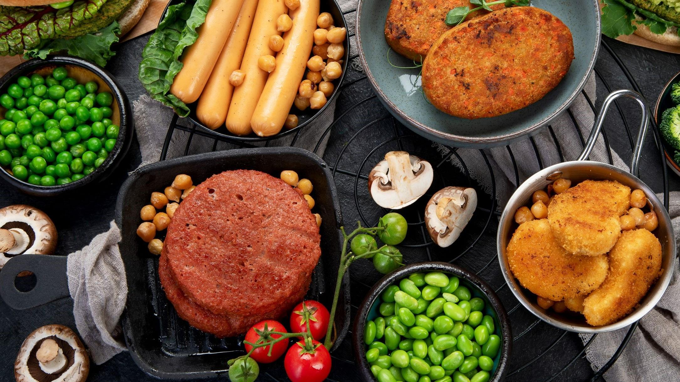 Ein Tisch voll Alternativer Proteine: Hülsenfrüchte, Pilze, Würstchen und Schnitzel aus Fleischersatz.